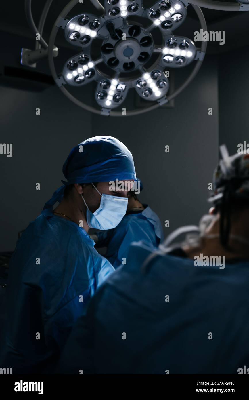 Portrait d'un médecin masculin en masque dans une chambre d'hôpital sombre avec équipement technologique. le chirurgien regarde le moniteur et opère le patient. Prise de vue verticale Banque D'Images