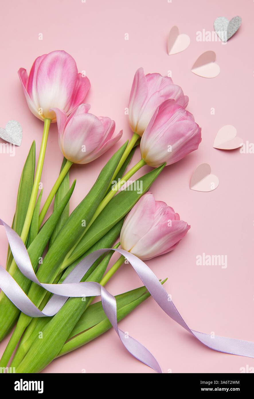 Beau bouquet de tulipes roses nouées avec un ruban violet couché sur une surface rose décorée de coeurs en papier, créant une belle composition printanière Banque D'Images