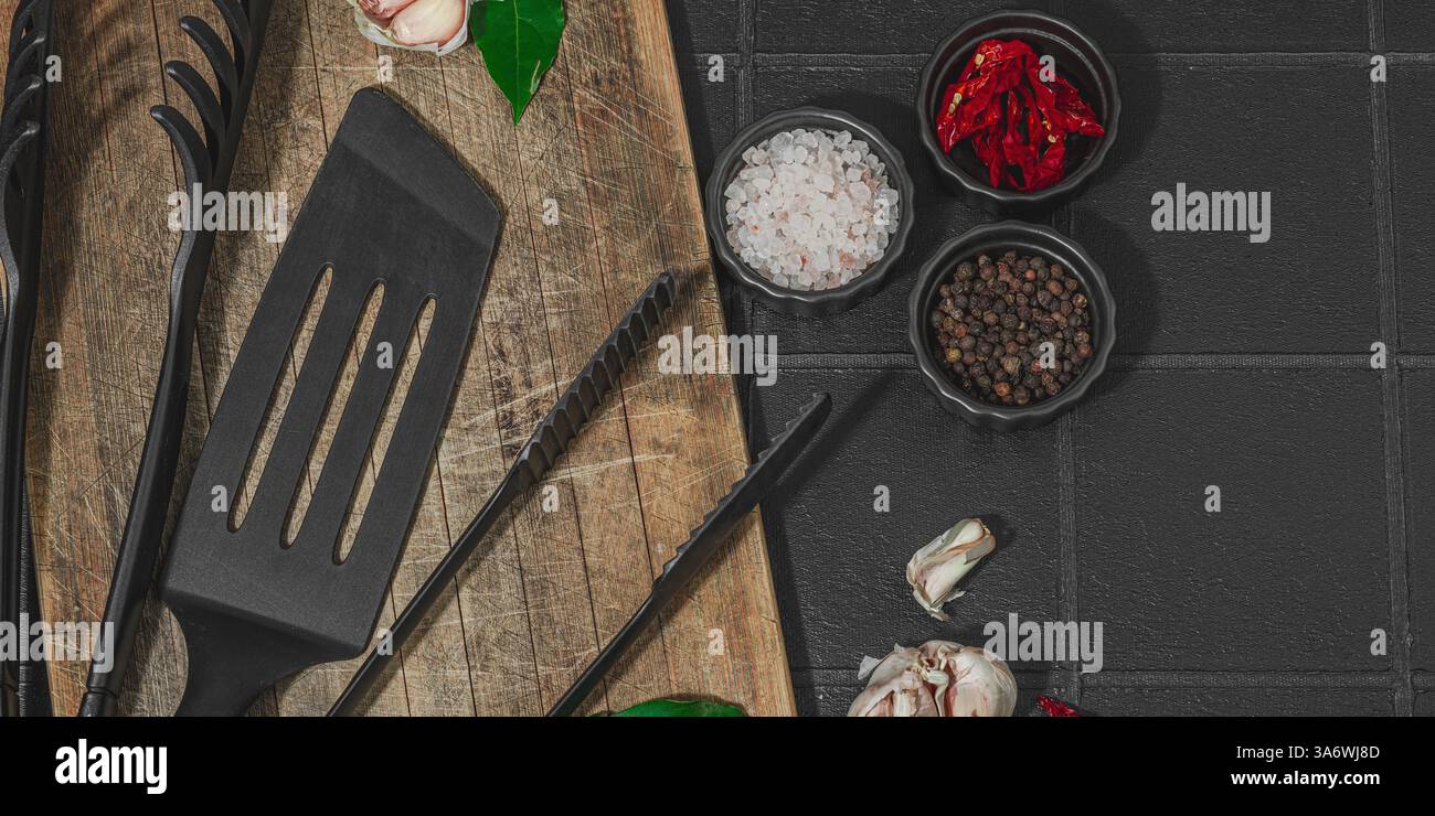 Concept de saison barbecue. Pinces à griller, spatule, set d'épices, gros sel, ail et herbes fraîches. Préparation des aliments grillés, fond de tuiles noires, plat Banque D'Images
