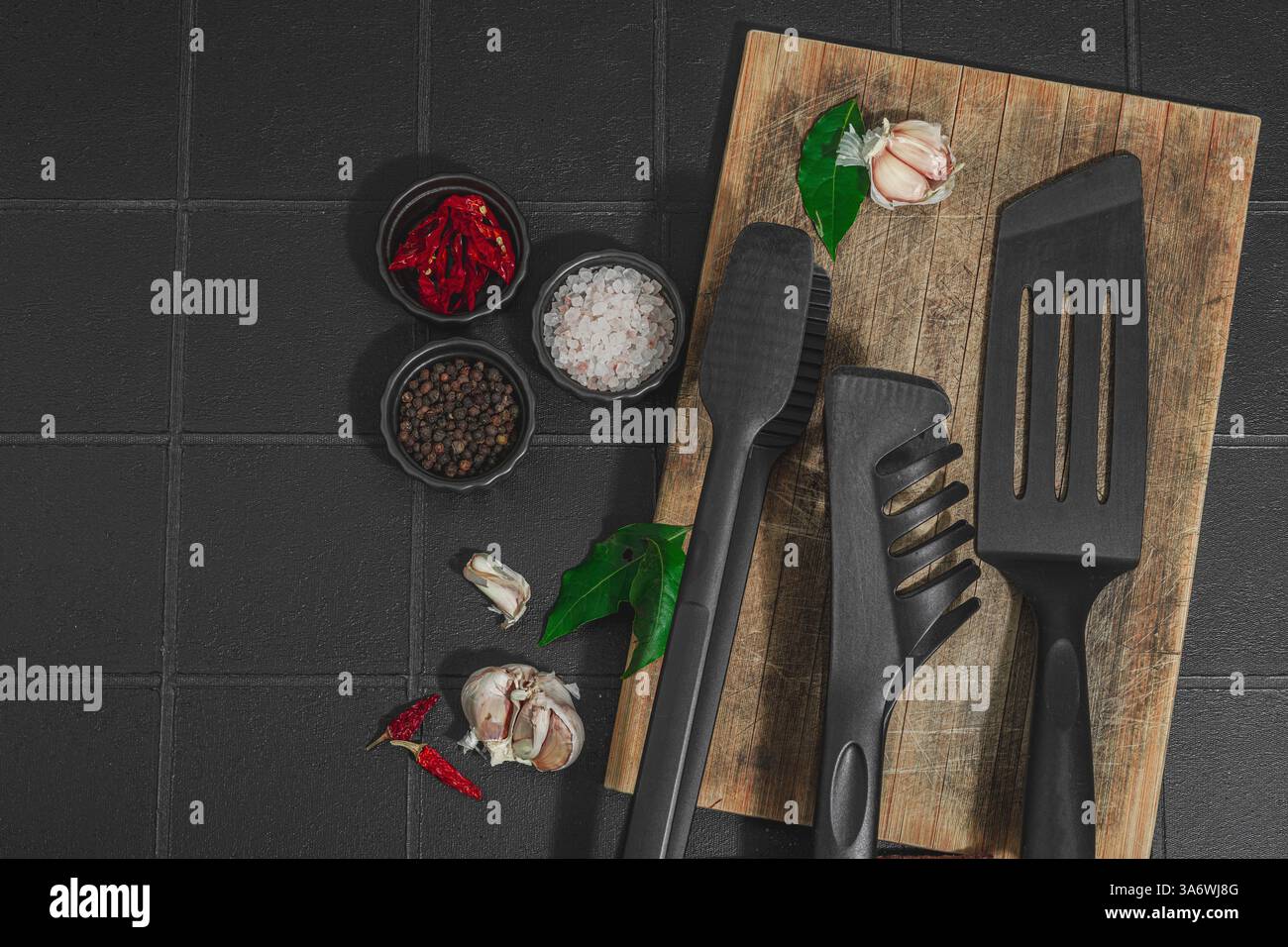 Concept de saison barbecue. Pinces à griller, spatule, set d'épices, gros sel, ail et herbes fraîches. Préparation des aliments grillés, fond de tuiles noires, plat Banque D'Images
