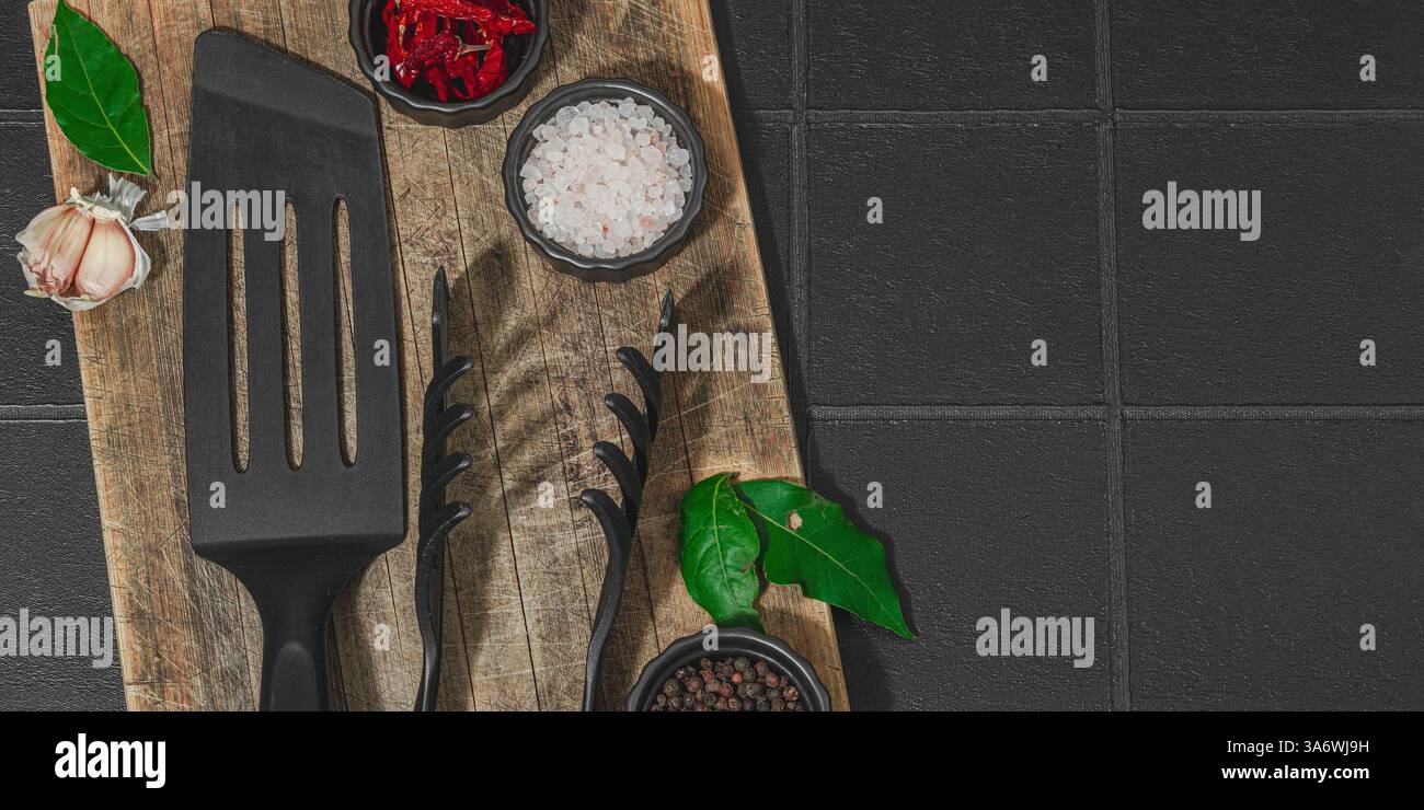 Concept de saison barbecue. Pinces à griller, spatule, set d'épices, gros sel, ail et herbes fraîches. Préparation des aliments grillés, fond de tuiles noires, plat Banque D'Images