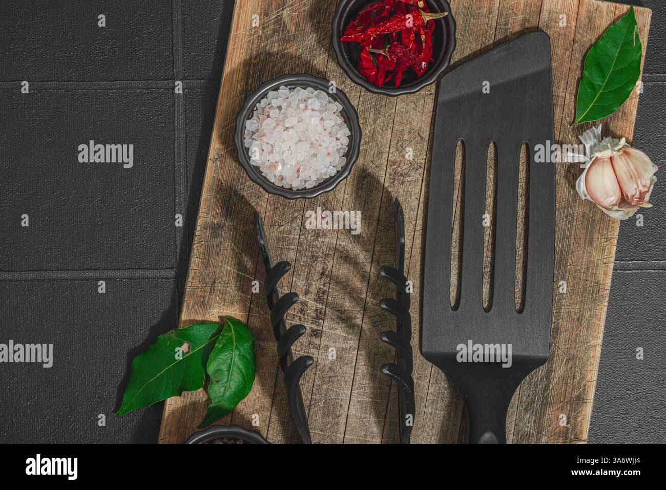 Concept de saison barbecue. Pinces à griller, spatule, set d'épices, gros sel, ail et herbes fraîches. Préparation des aliments grillés, fond de tuiles noires, plat Banque D'Images
