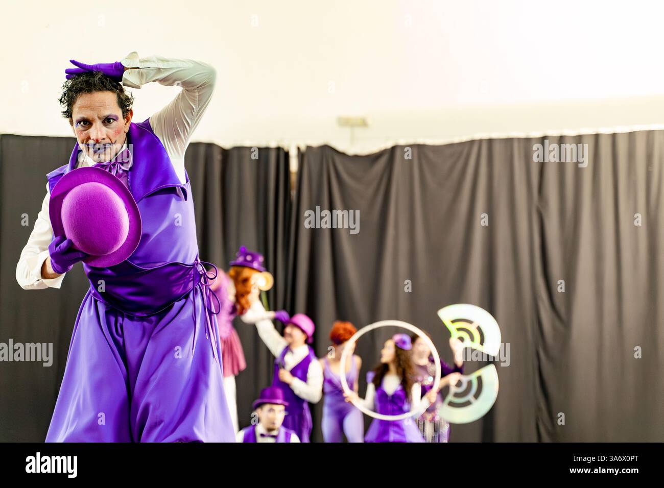 Clown masculin portant un costume violet et tenant un chapeau melon violet se produisant sur scène avec d'autres artistes flous en arrière-plan Banque D'Images