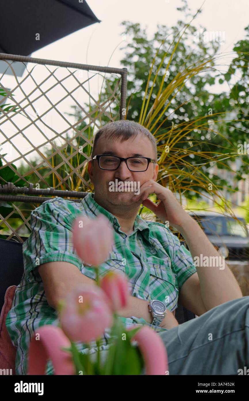 Un homme se détend sur une chaise de patio, entouré de plantes vertes luxuriantes et de tulipes roses vibrantes. Banque D'Images