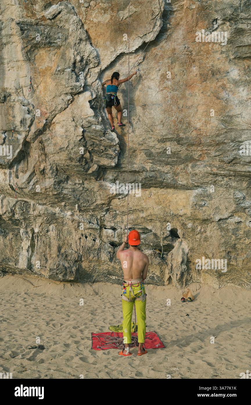 Grimpeur monte sur le mur de calcaire texturé à Tonsai tandis que son partenaire s'éloigne d'en bas Banque D'Images