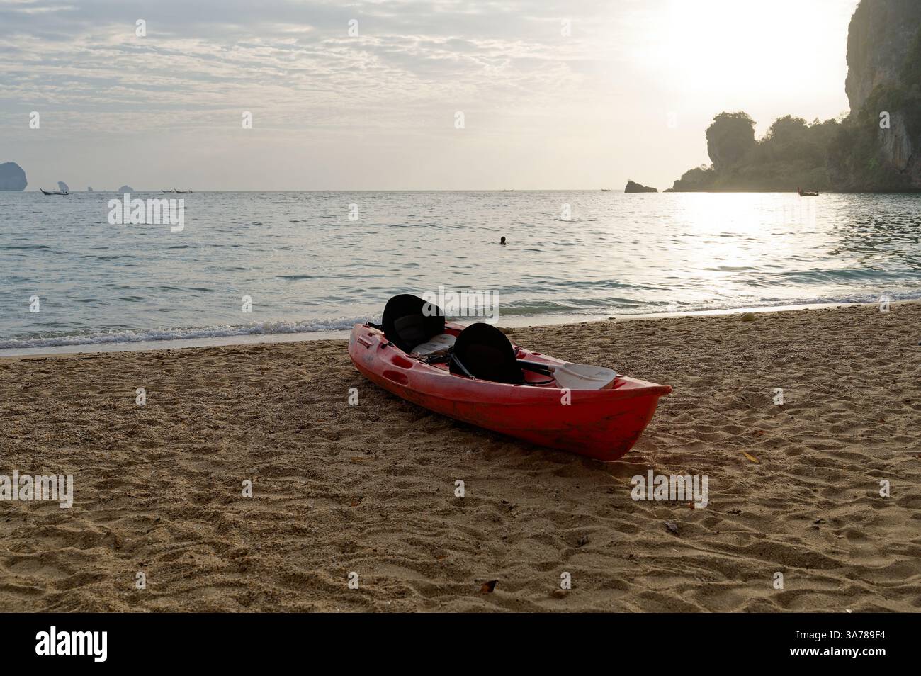Un kayak rouge seul repose sur la rive de la plage de Tonsai, baigné dans la douce lumière dorée du soleil couchant tandis qu'un nageur flotte paisiblement à proximité Banque D'Images