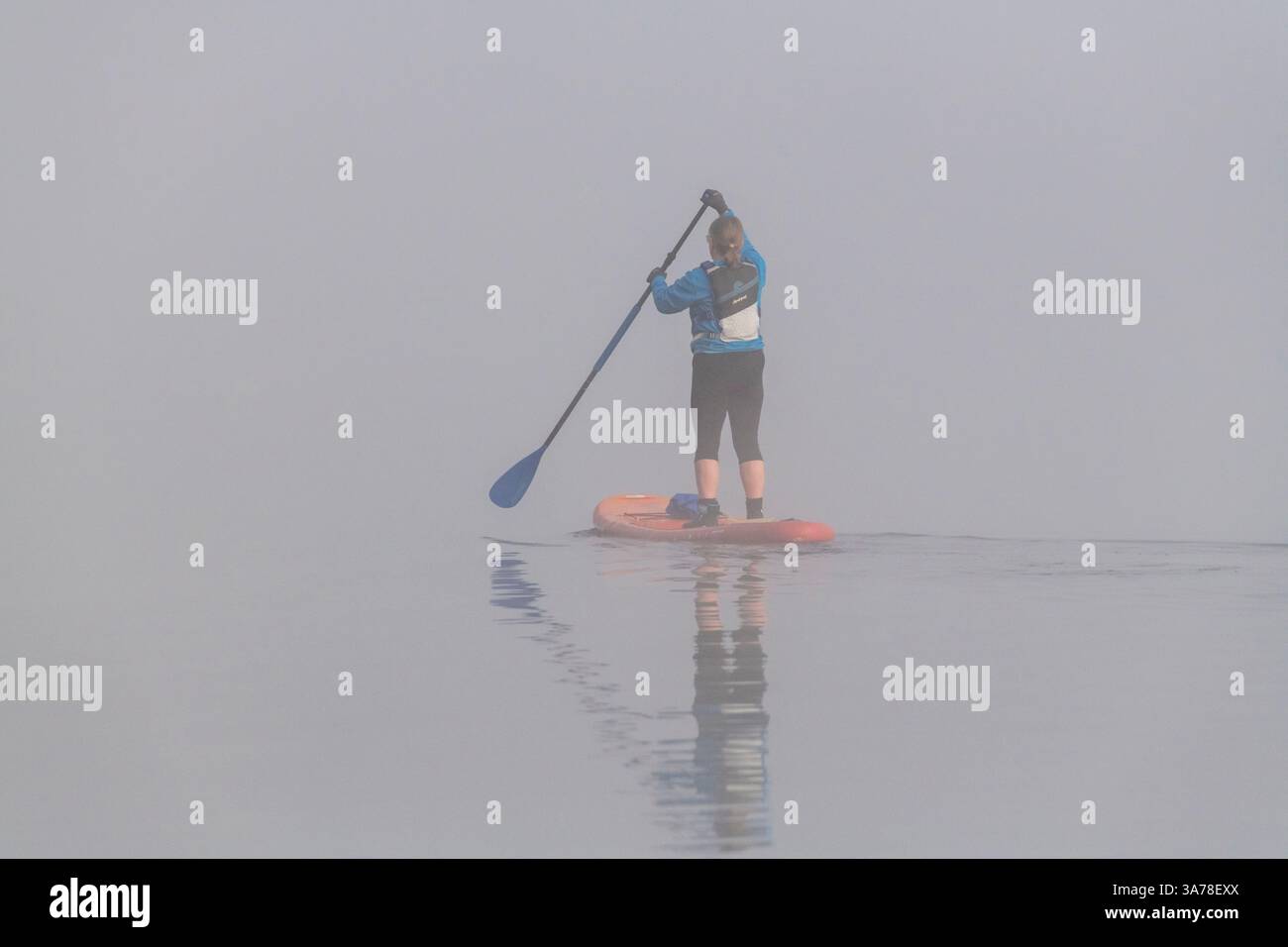 Une femme sur une planche de stand up paddle disparaissant dans le brouillard un matin calme au début du printemps Banque D'Images