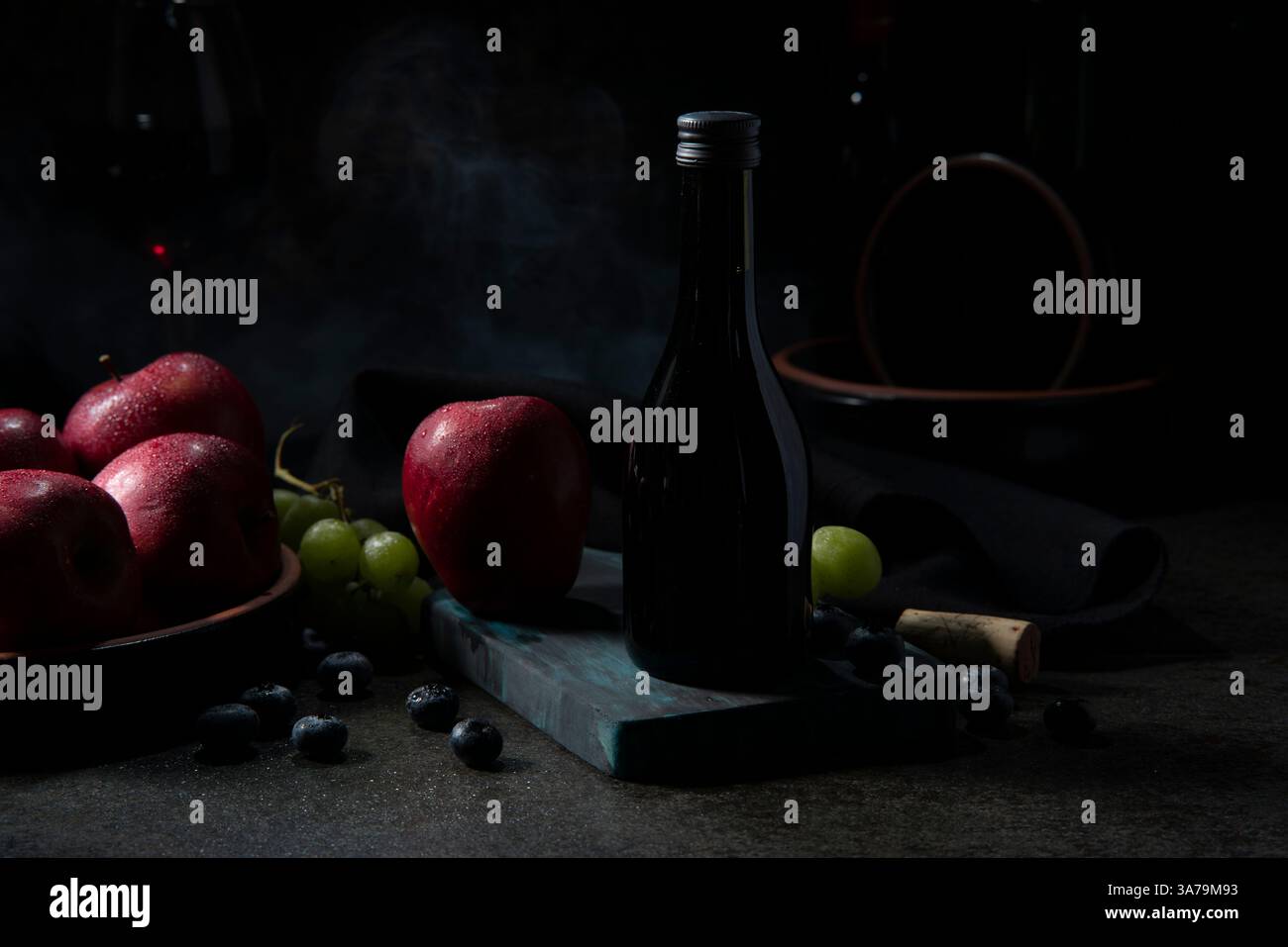 Bouteille de vin à côté des pommes rouges, des raisins verts et des myrtilles sur une planche à découper sur une table en céramique noire avec un tissu noir en arrière-plan Banque D'Images