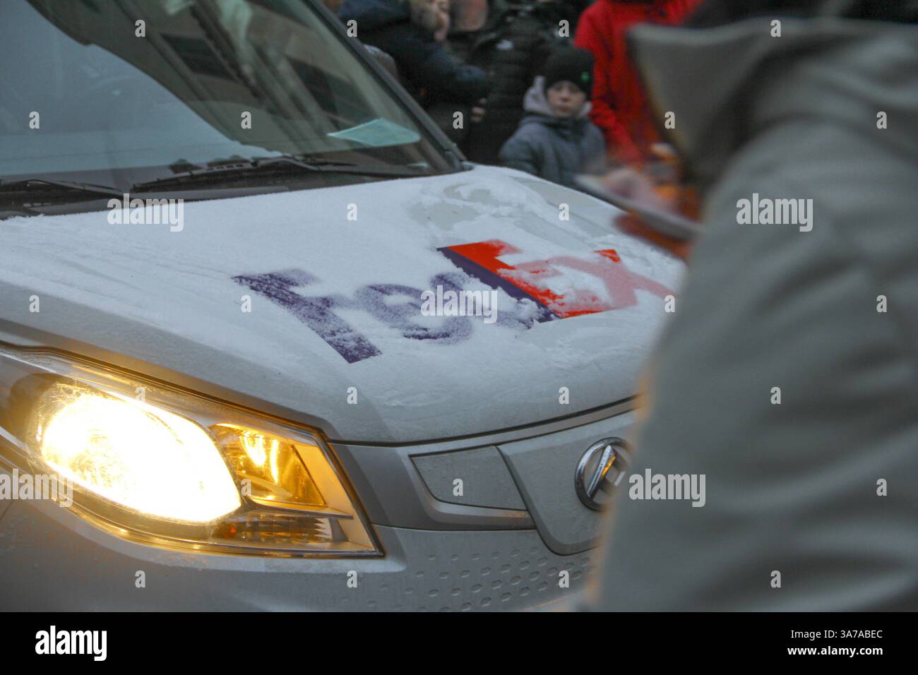 Close-up d'une camionnette de livraison FedEx givrée avec des phares allumés dans un cadre urbain enneigé, avec des personnes floues en vêtements d'hiver en arrière-plan. Banque D'Images