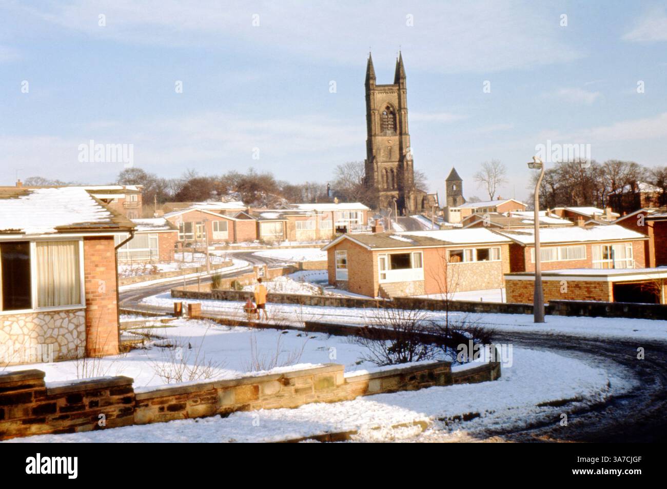 Photographiée en avril 1969, cette image montre une vue enneigée de la banlieue de Flash Lane à Mirfield, West Yorkshire. La scène capture un logement du milieu du siècle niché dans le paysage hivernal, avec l'importante église jumelée de Marie s'élevant en arrière-plan. Une figure solitaire marche le long de la rue sinueuse. Banque D'Images