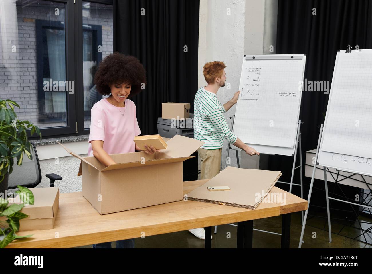 Dans un bureau contemporain, une personne déballe joyeusement une boîte tandis qu’une autre prépare une présentation. Banque D'Images