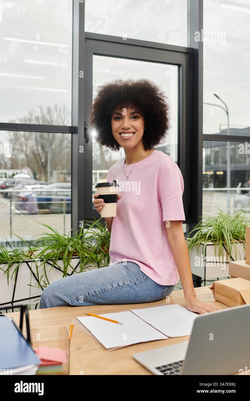 Une femme joyeuse sirote un café à une table, entourée de matériaux d'emballage dans un espace de travail moderne. Banque D'Images