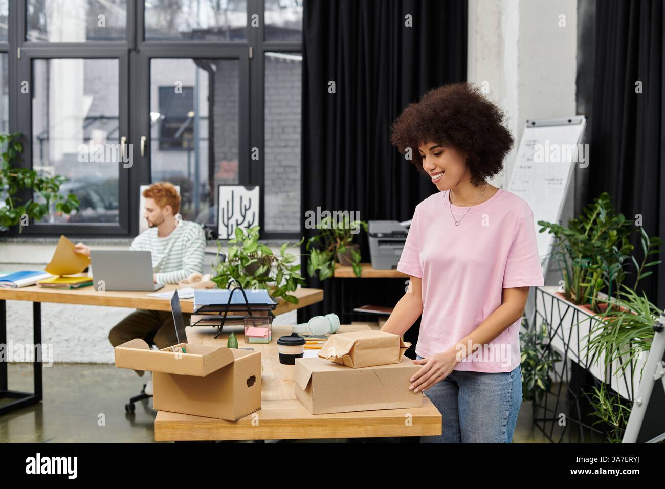 Une personne déballe des boîtes dans un espace de travail lumineux et rempli de plantes tandis qu'une autre se concentre. Banque D'Images