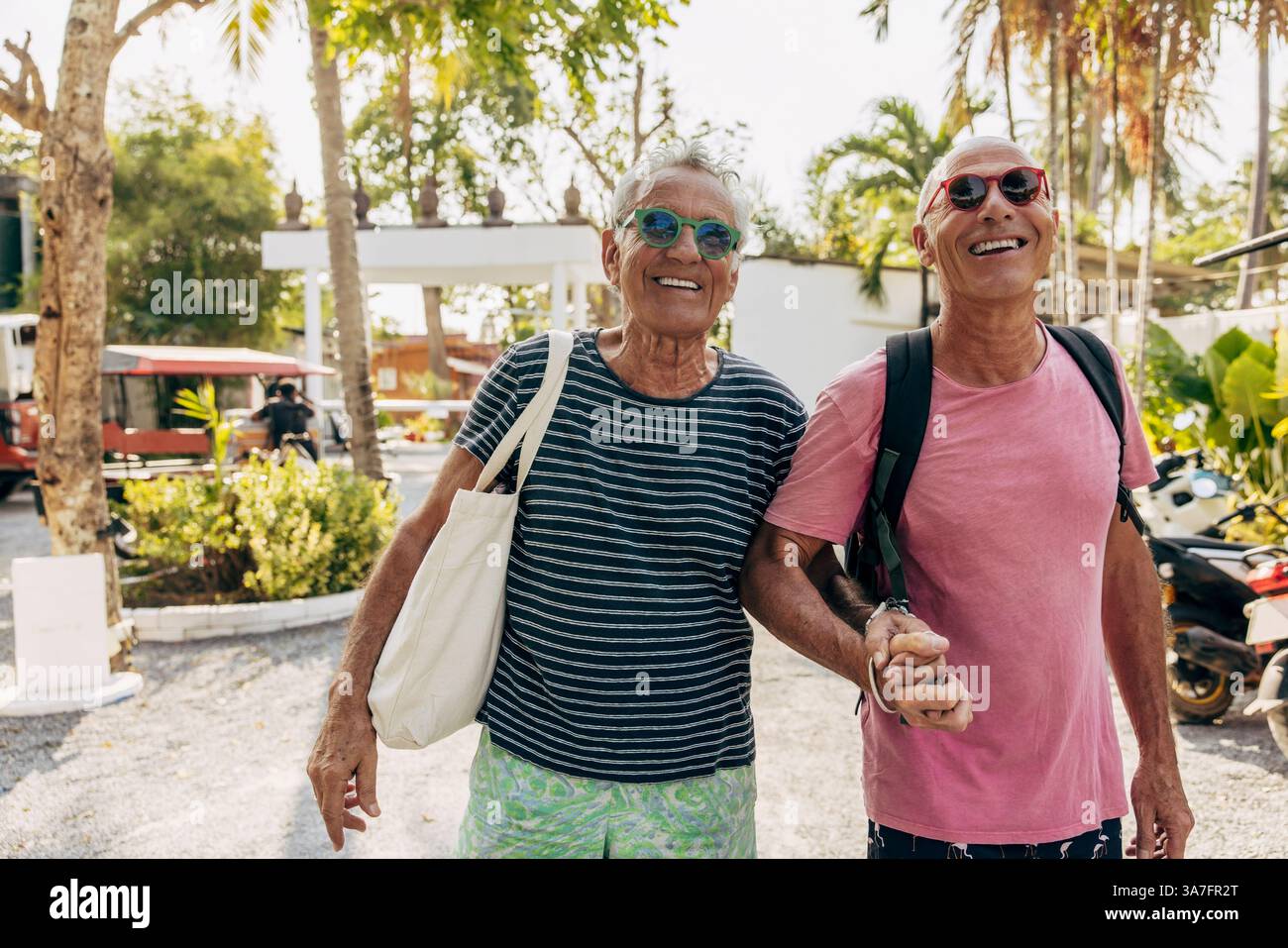 Heureux couple homosexuel âgé portant des lunettes de soleil se tenant la main tout en marchant à destination de vacances Banque D'Images