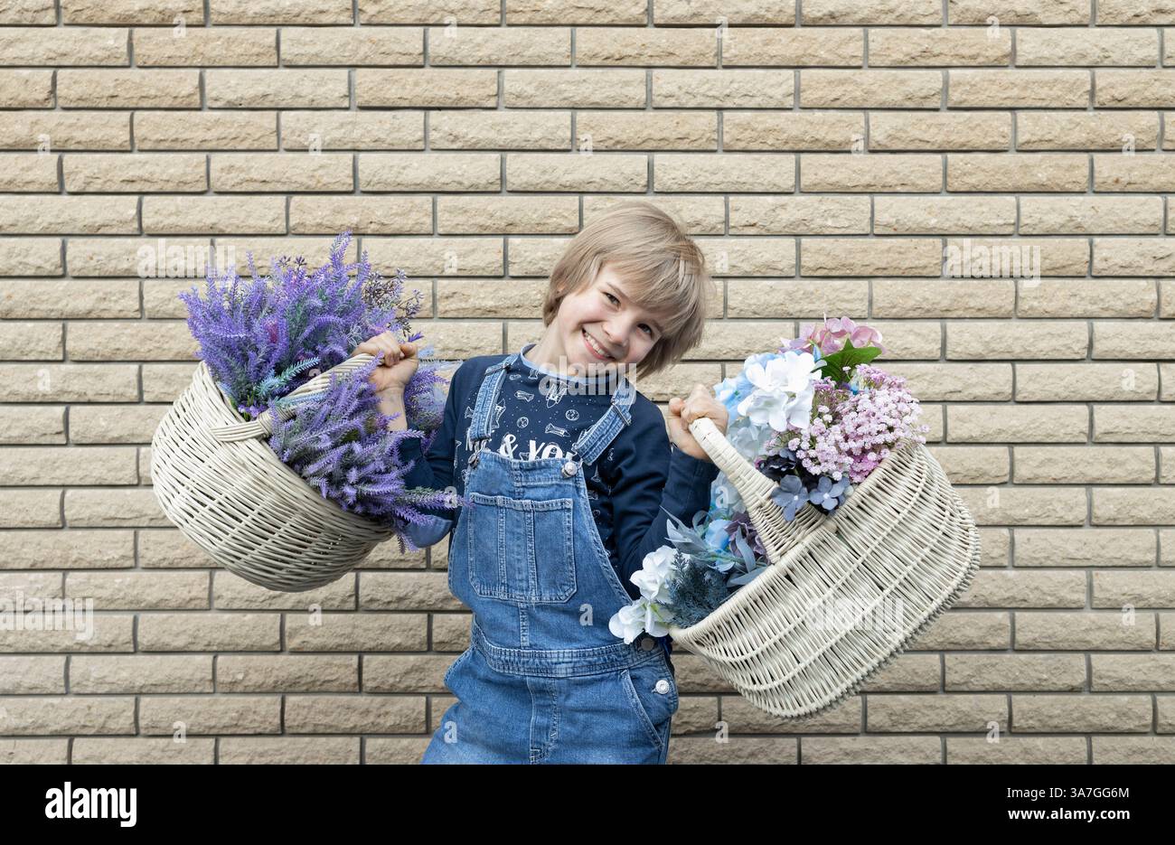 Cute Happy Boy de 8 ans en combinaison en denim tient deux grands paniers en osier avec des fleurs sur fond de mur de briques. Concept de livraison de fleurs, chil souriant Banque D'Images