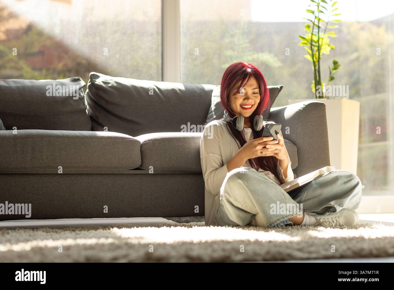 Femme asiatique heureuse avec des écouteurs assis sur un tapis et utilisant un smartphone Banque D'Images
