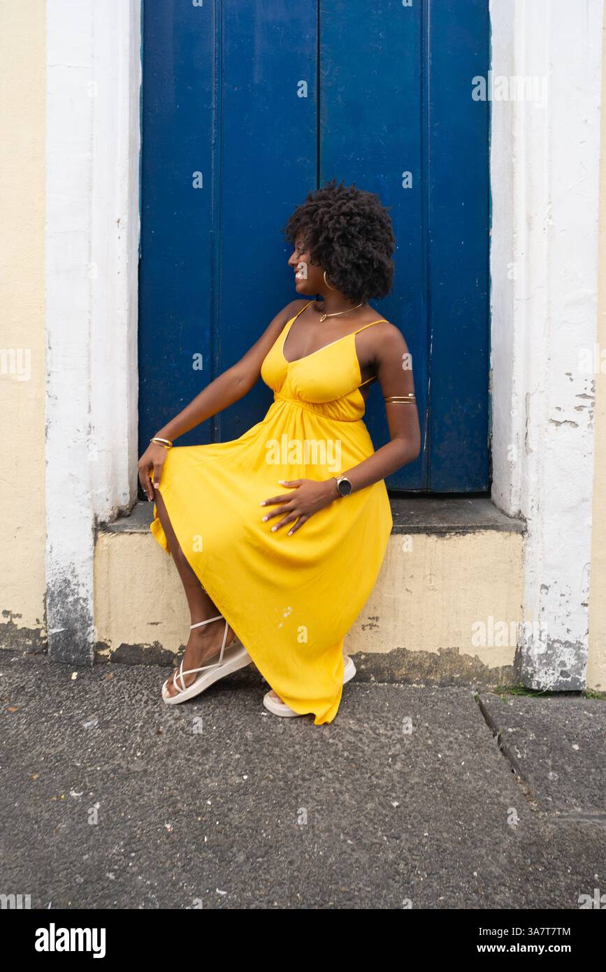 Belle jeune femme aux cheveux afro, vêtue d'une robe jaune, assise près d'une porte bleue d'une vieille maison. Personne confiante. Brésil Banque D'Images