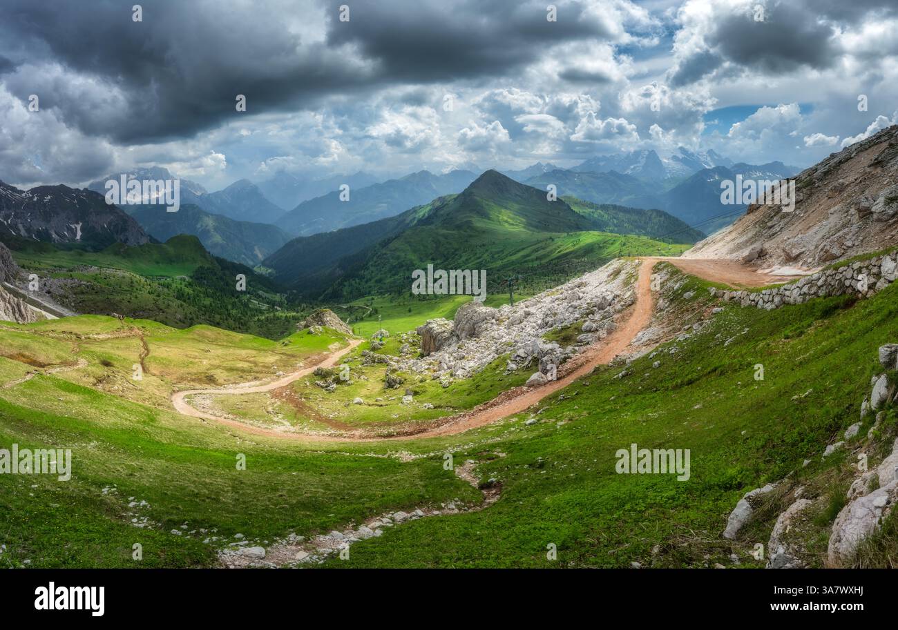 Route de montagne sinueuse dans une vallée verdoyante, les sommets alpins sous un ciel spectaculaire rempli de lumière et d'ombre contrastées en été. Panora Banque D'Images