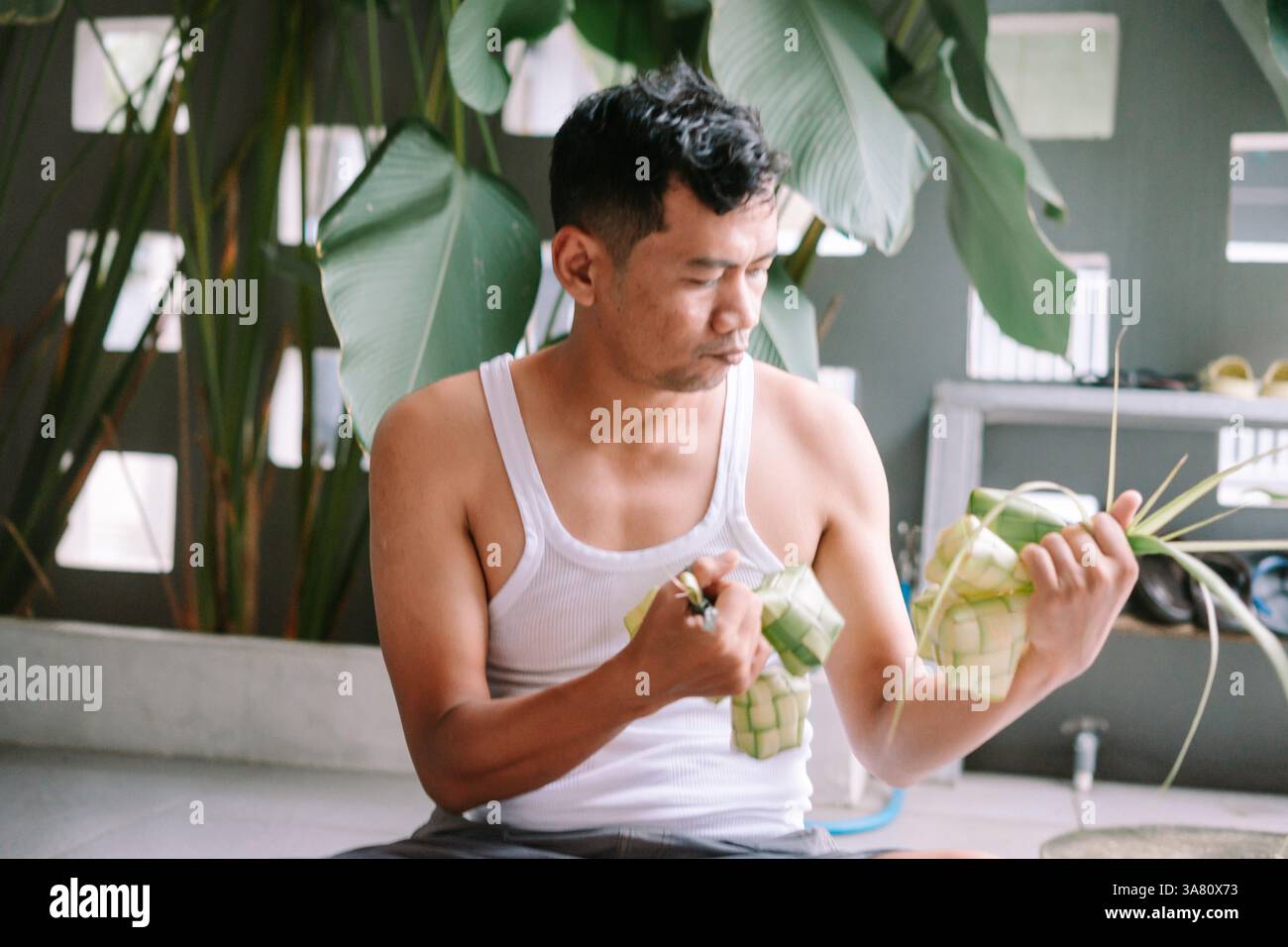 Un homme asiatique fabrique habilement du ketupat, une boulette de riz traditionnelle en feuilles de palmier tissées, se préparant à une célébration culturelle. Il démontre l'un Banque D'Images