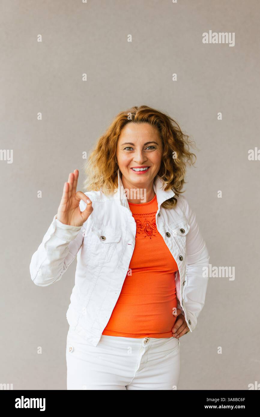 Joyeuse femme d'âge moyen dans un haut orange et une veste blanche faisant un geste correct avec sa main tout en souriant avec confiance sur un fond Uni Banque D'Images