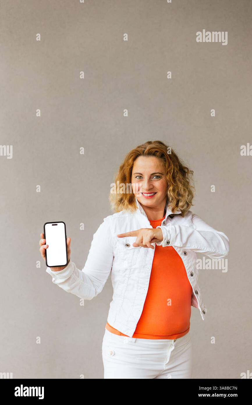 Femme d'âge moyen souriante en haut orange et veste blanche tenant un smartphone avec un écran vierge et pointant vers lui, debout contre un dos neutre Banque D'Images