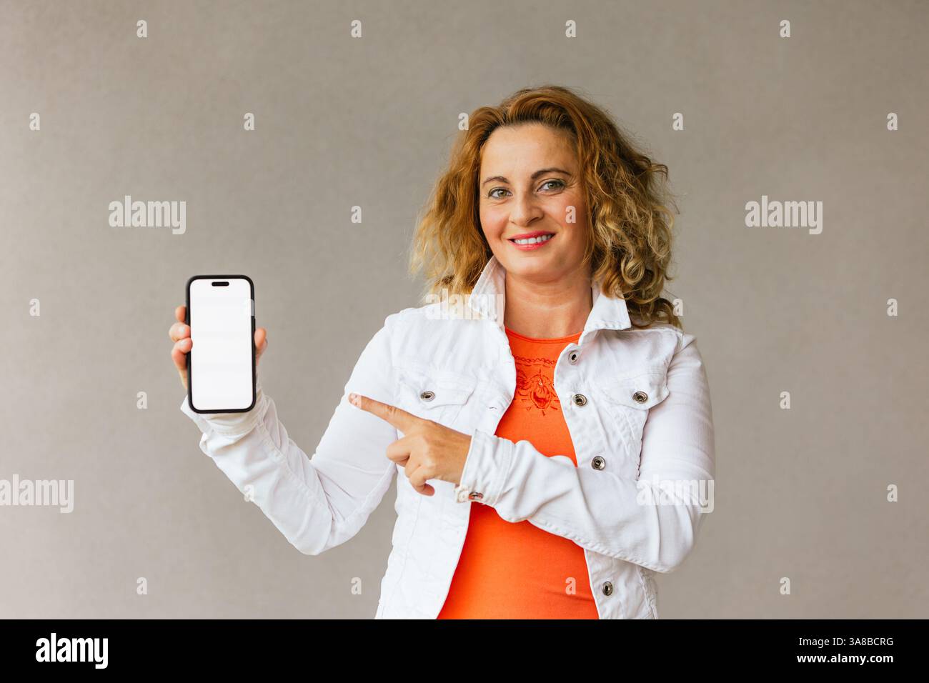 Femme d'âge moyen dans un haut orange et une veste blanche tenant un smartphone avec écran vierge et pointant vers lui, promouvant l'application mobile ou le contenu avec co Banque D'Images