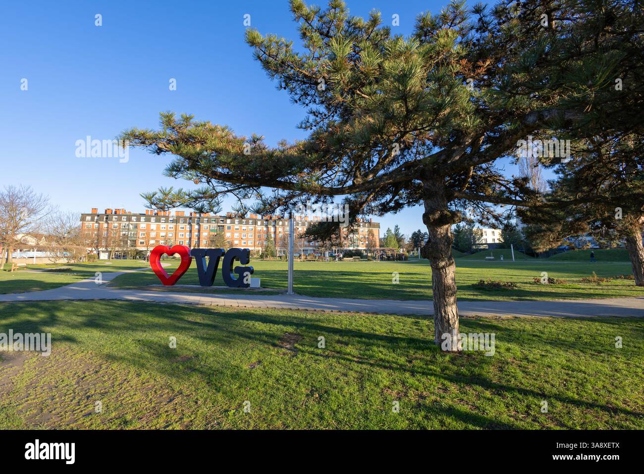 Un parc urbain lumineux avec un pin, une herbe bien entretenue et une installation animée sur le thème de l'amour, avec des bâtiments en arrière-plan sous un blu Banque D'Images