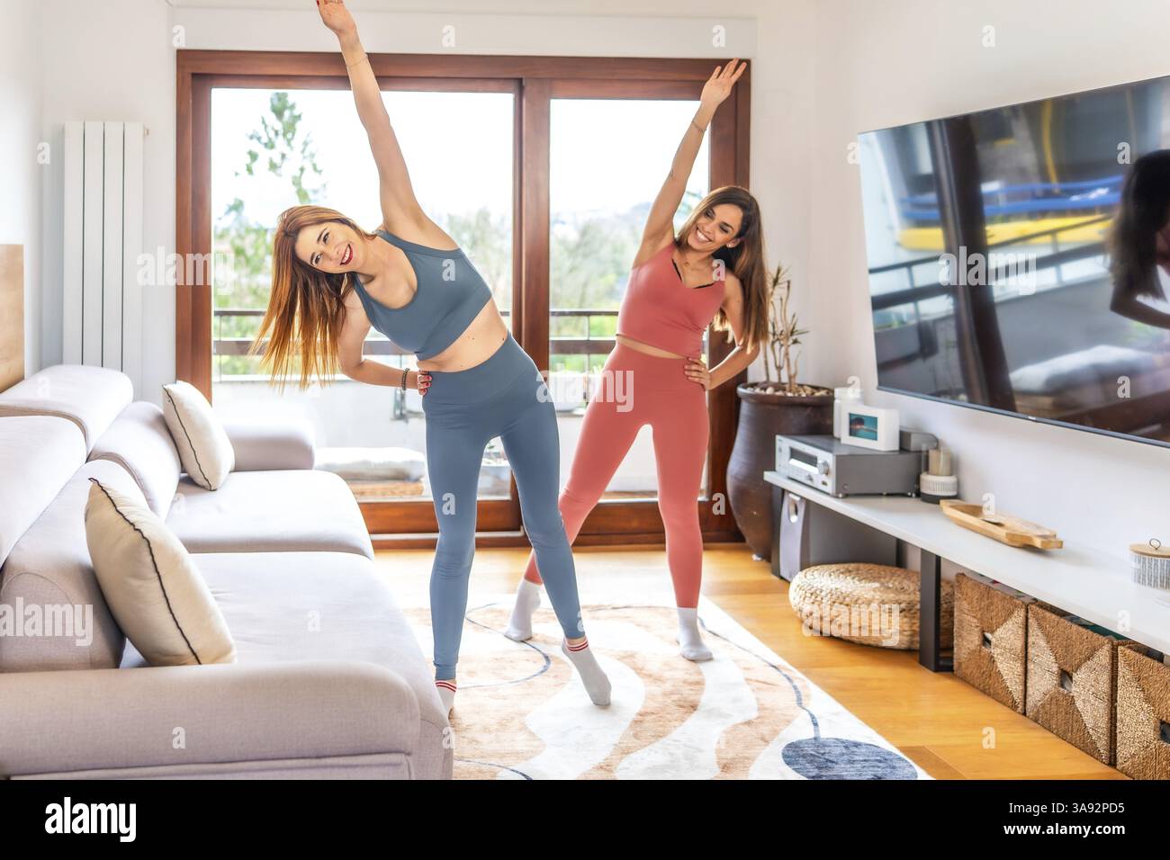Deux femmes souriantes pratiquant le yoga ensemble dans leur salon, suite à un cours en ligne sur leur télé Banque D'Images