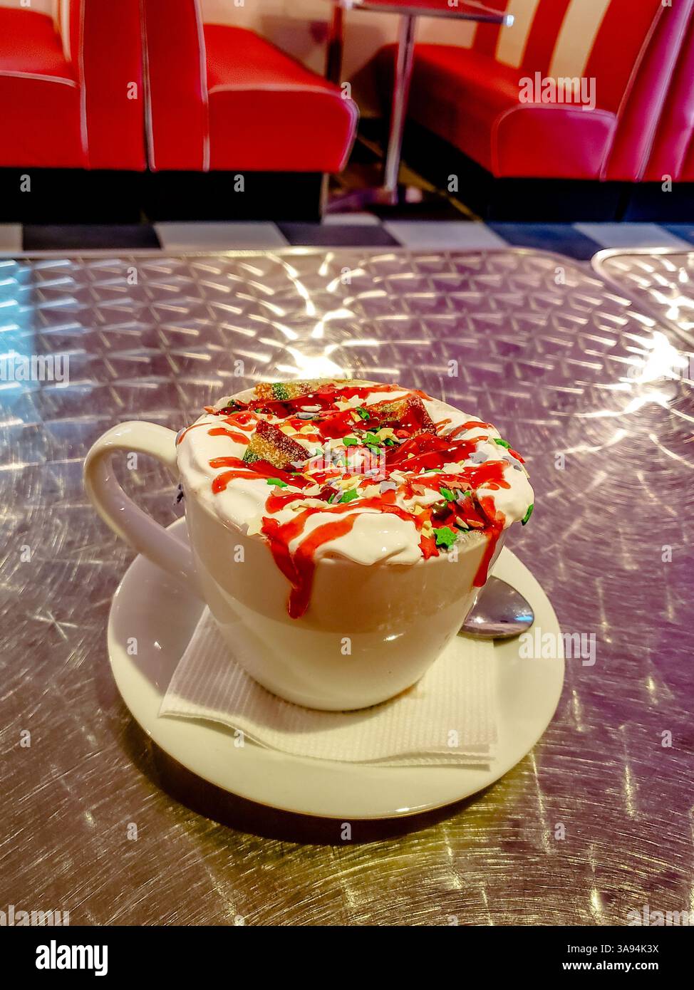Café festif à la crème fouettée, sirop rouge et saupoudres servi dans une tasse blanche sur soucoupe dans un restaurant américain rétro avec table en métal brillant Banque D'Images