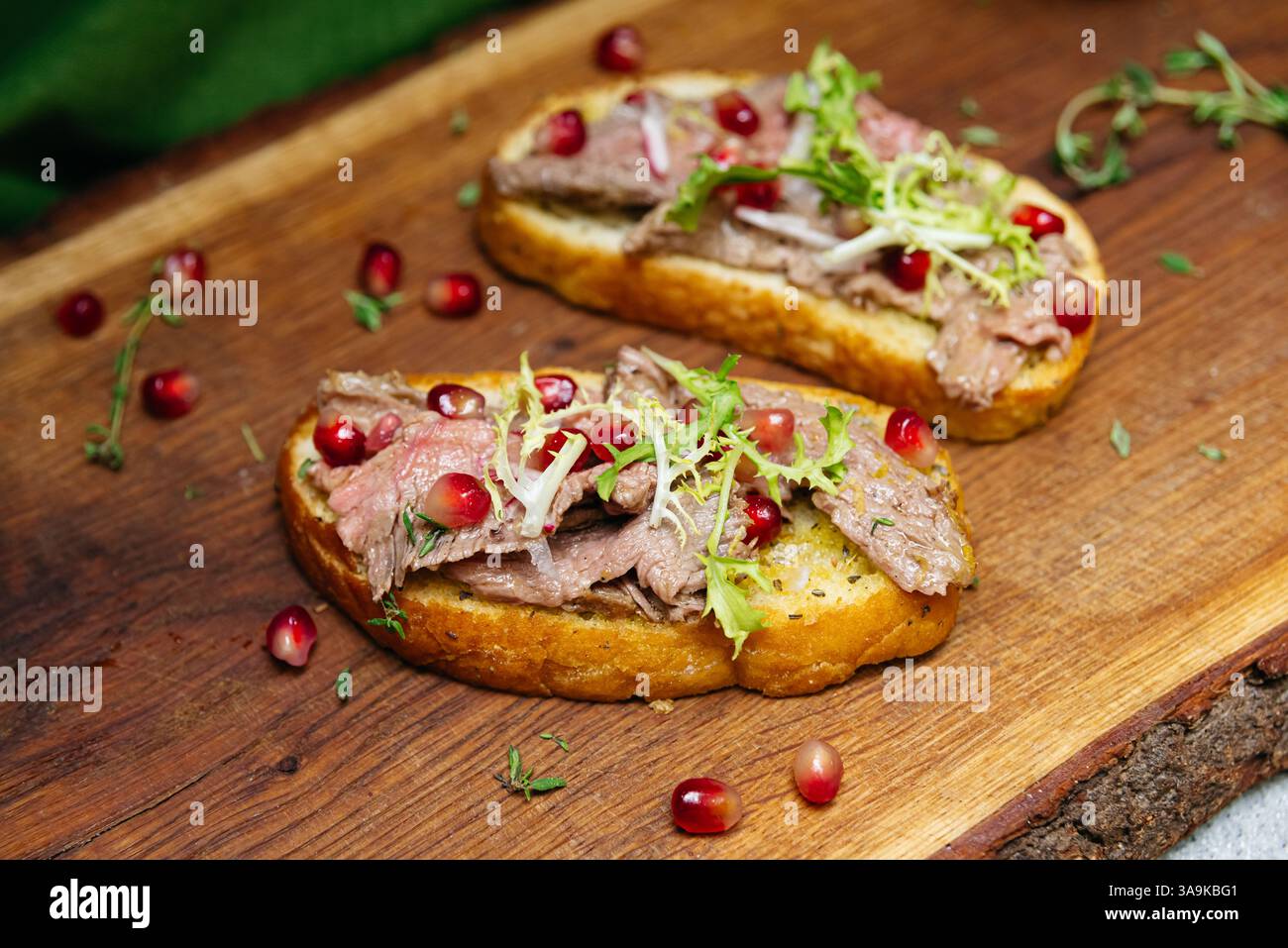 Pain grillé garni de tranches de bœuf, d'herbes fraîches et de graines de grenade servi sur une planche de bois rustique pour une présentation gastronomique. Banque D'Images
