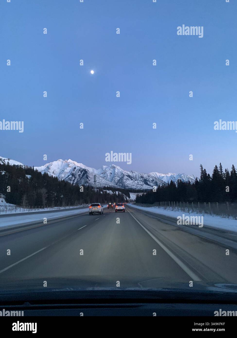 Route panoramique vers des montagnes enneigées sous un ciel crépusculaire, avec la lune se levant au-dessus des Rocheuses canadiennes. Un superbe Road trip hivernal. - Image de stock capturée avec un smartphone