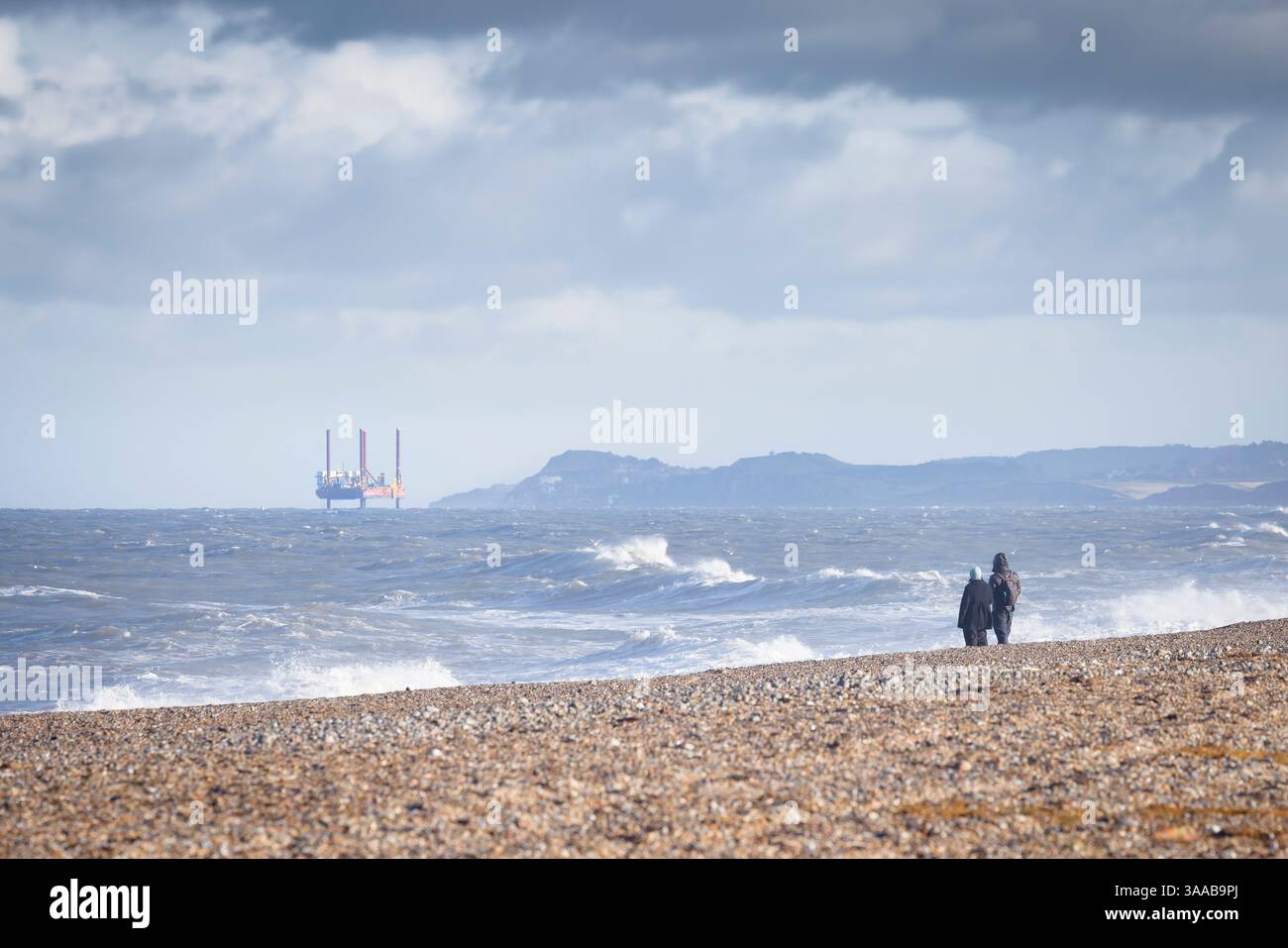 Les gens sur la plage de galets en hiver, regardant une plate-forme pétrolière de levage lointaine dans la mer du Nord. Blakeney point, Norfolk, Royaume-Uni Banque D'Images