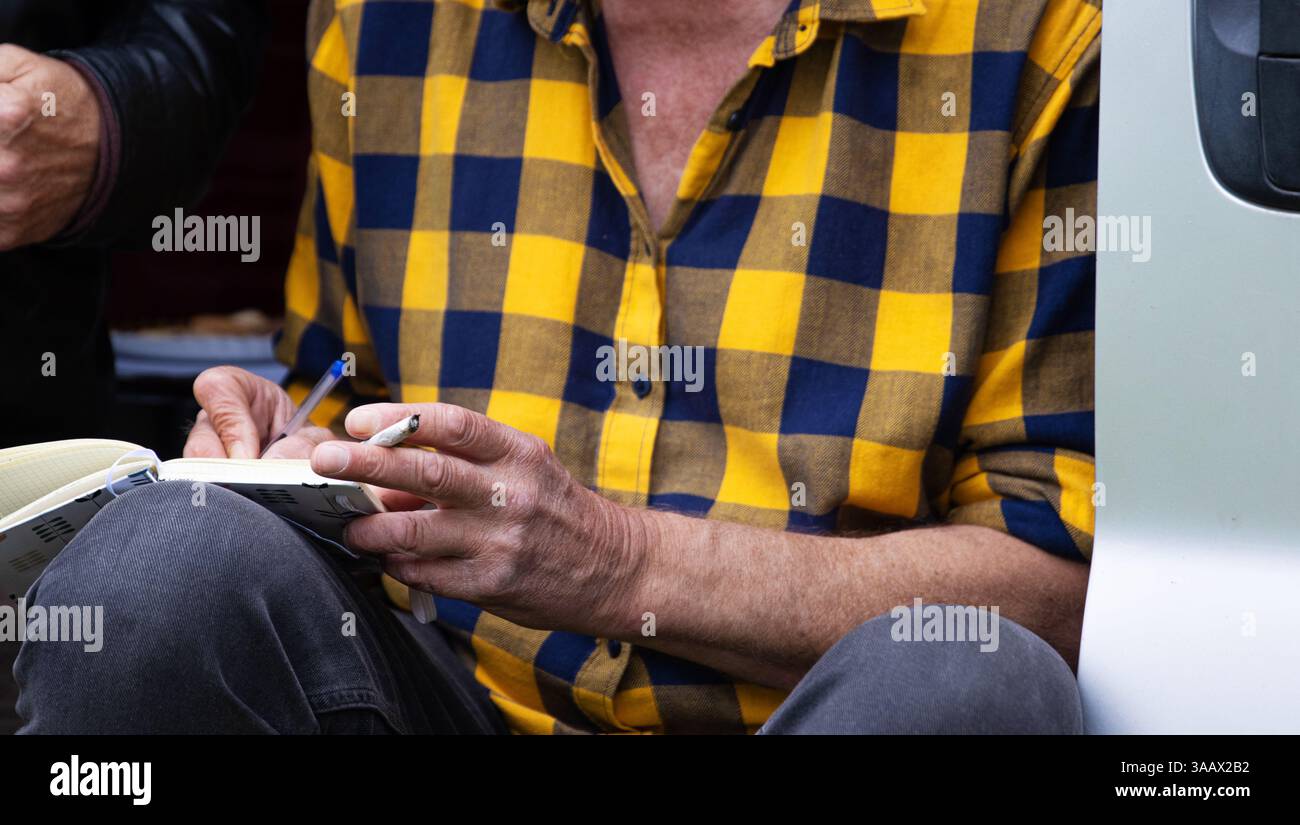 Gros plan d'un homme dans une chemise jaune à carreaux tenant un stylo et une cigarette, assis im camping-car van et écrivant dans le carnet habitudes de fumer, créativité, Banque D'Images