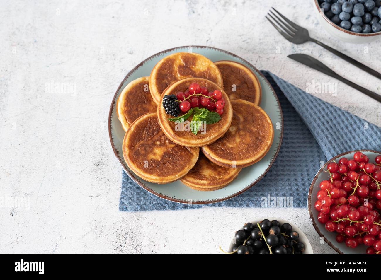 Pile de crêpes maison avec des bleuets, des framboises et des fraises sur une assiette blanche avec un chiffon bleu sur un dessus de table gris clair Banque D'Images Pile de crêpes maison avec des bleuets, des framboises et des fraises sur une assiette blanche avec un chiffon bleu sur un dessus de table gris clair Banque D'Images