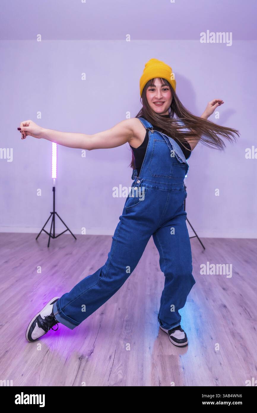 Danseuse portant une combinaison en denim et un bonnet jaune présentant des mouvements de danse énergiques dans un studio éclairé par des lumières LED vibrantes Banque D'Images