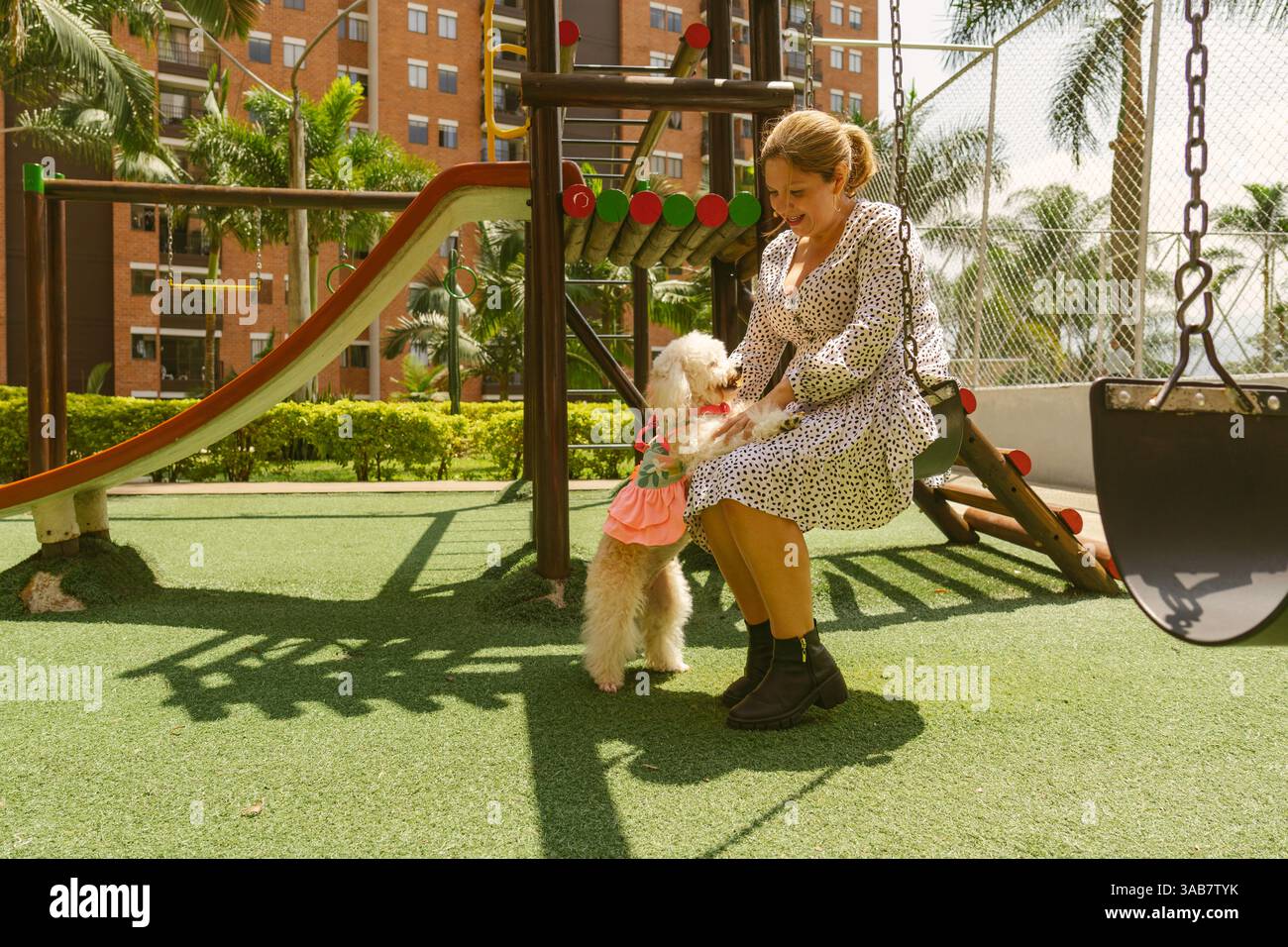 Femme heureuse dans une robe élégante balançant sur une balançoire de terrain de jeu, jouant joyeusement avec son petit chien blanc habillé en rose, créant des moments de plaisir et Banque D'Images