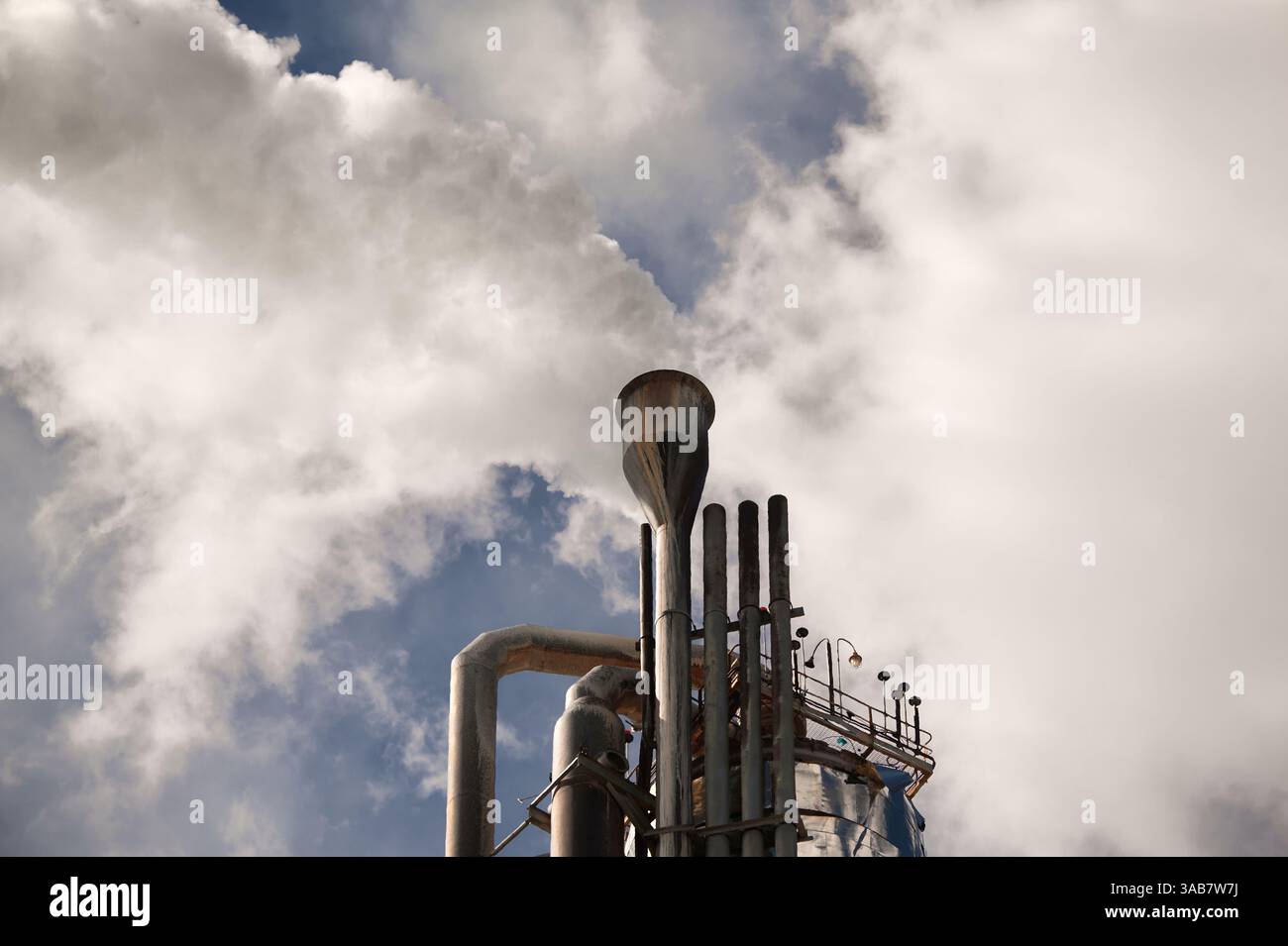 Colonne de distillation dans une usine chimique libérant de la vapeur sous un ciel bleu nuageux dans une zone industrielle animée pendant la journée Banque D'Images