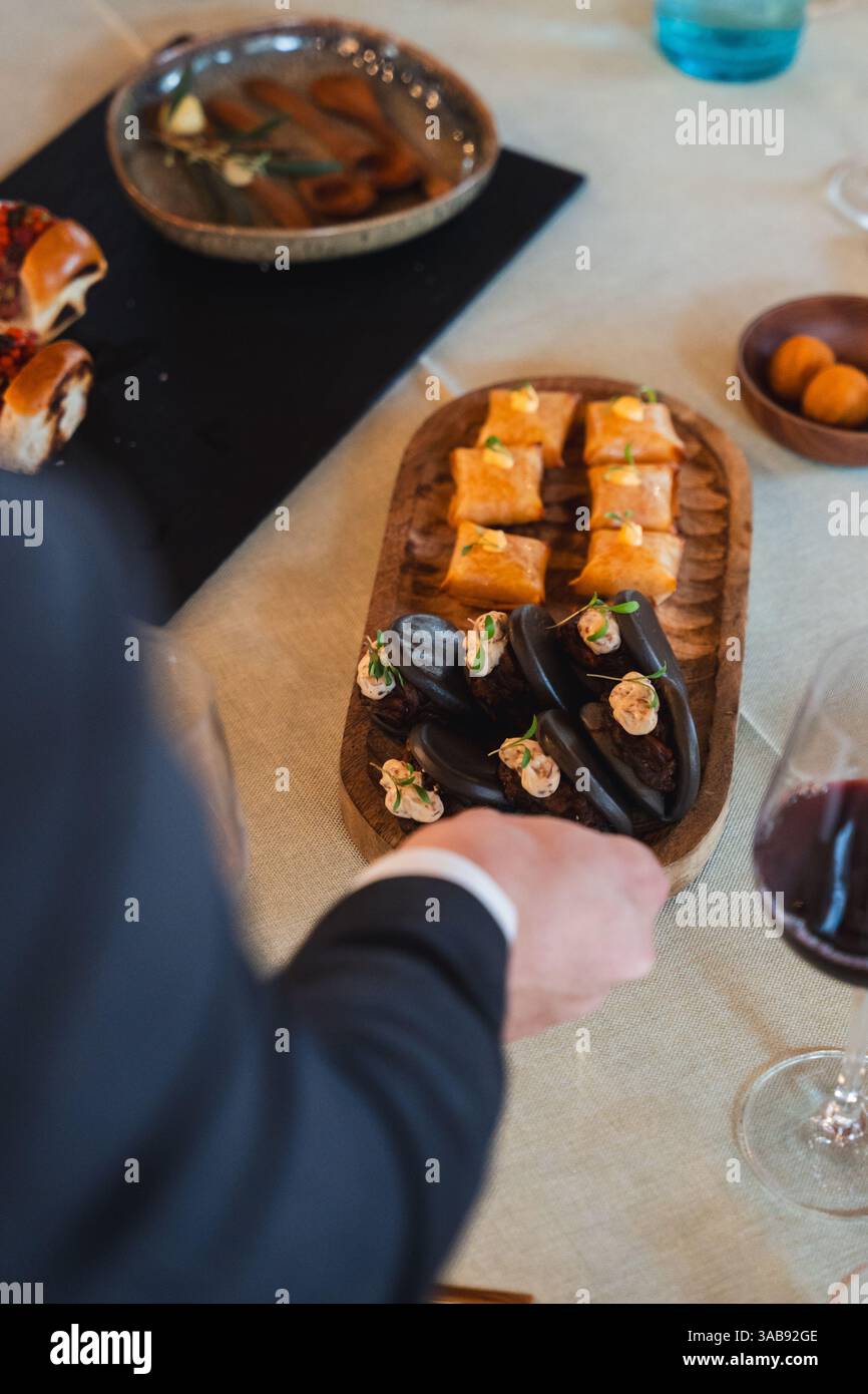 Un invité en tenue habillée va chercher un plateau en bois magnifiquement arrangé rempli de petits pains bao au poulpe et de hors-d'œuvre brun doré, sur une étiquette raffinée Banque D'Images
