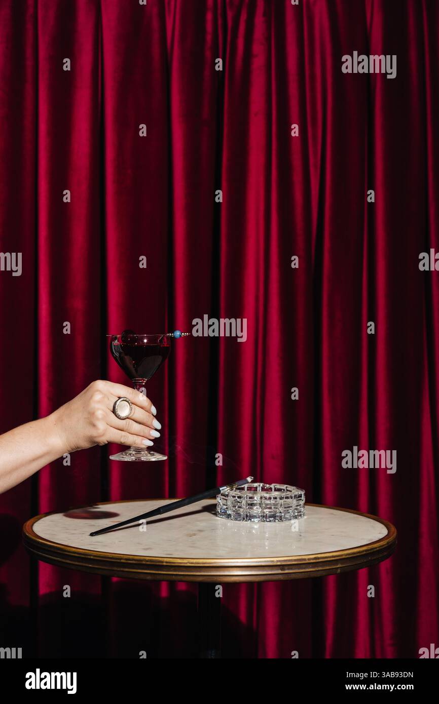 Une main élégante tenant un cocktail de gin infusé de baies avec des notes florales et des éclaboussures de vin mousseux. Ensemble contre de riches rideaux de velours rouge, un tabl en marbre Banque D'Images