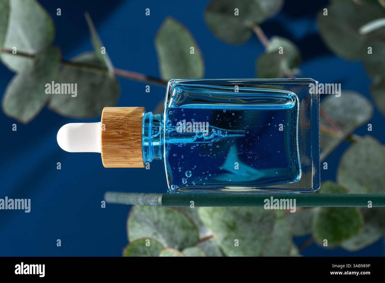 Un sérum ou une huile bleu visage conçu pour les hommes, contenu dans un flacon compte-gouttes en verre transparent avec un bouchon en bois naturel, placé sur un fond bleu foncé avec Banque D'Images