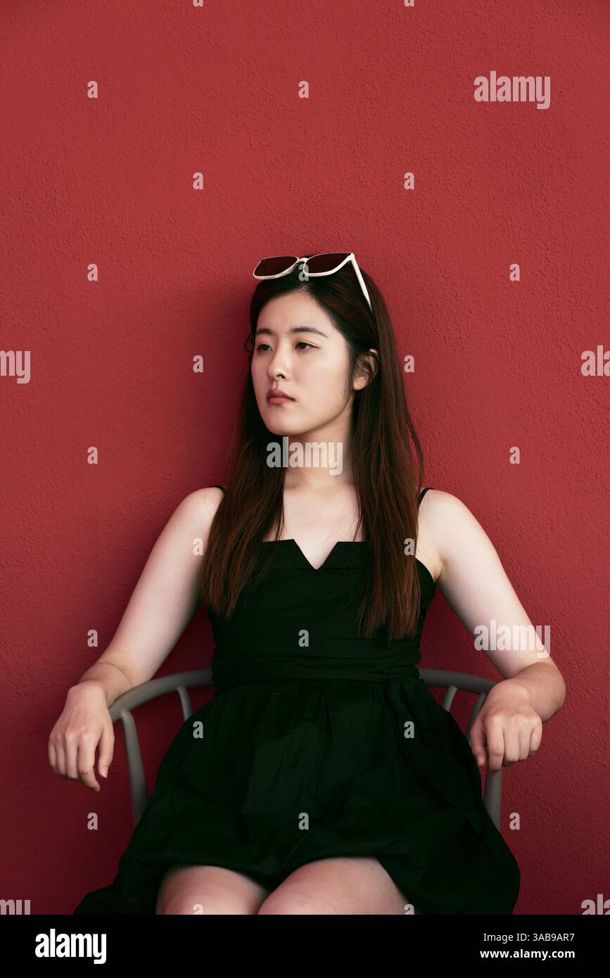 Une jeune femme asiatique dans une robe noire et des lunettes de soleil est assise contre un mur rouge vif. Son attitude calme et les couleurs contrastées créent un vi frappant Banque D'Images