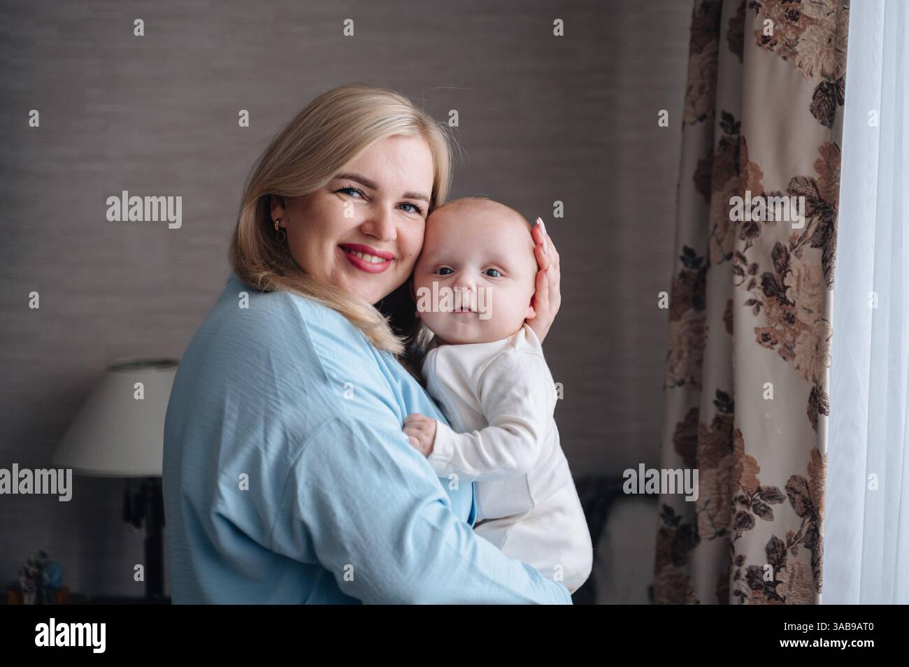 Une mère joyeuse tient amoureusement son fils bébé près d'une fenêtre, enveloppée d'une douce lumière naturelle. Le cadre serein de la maison rehausse ce moment de chaleur a Banque D'Images