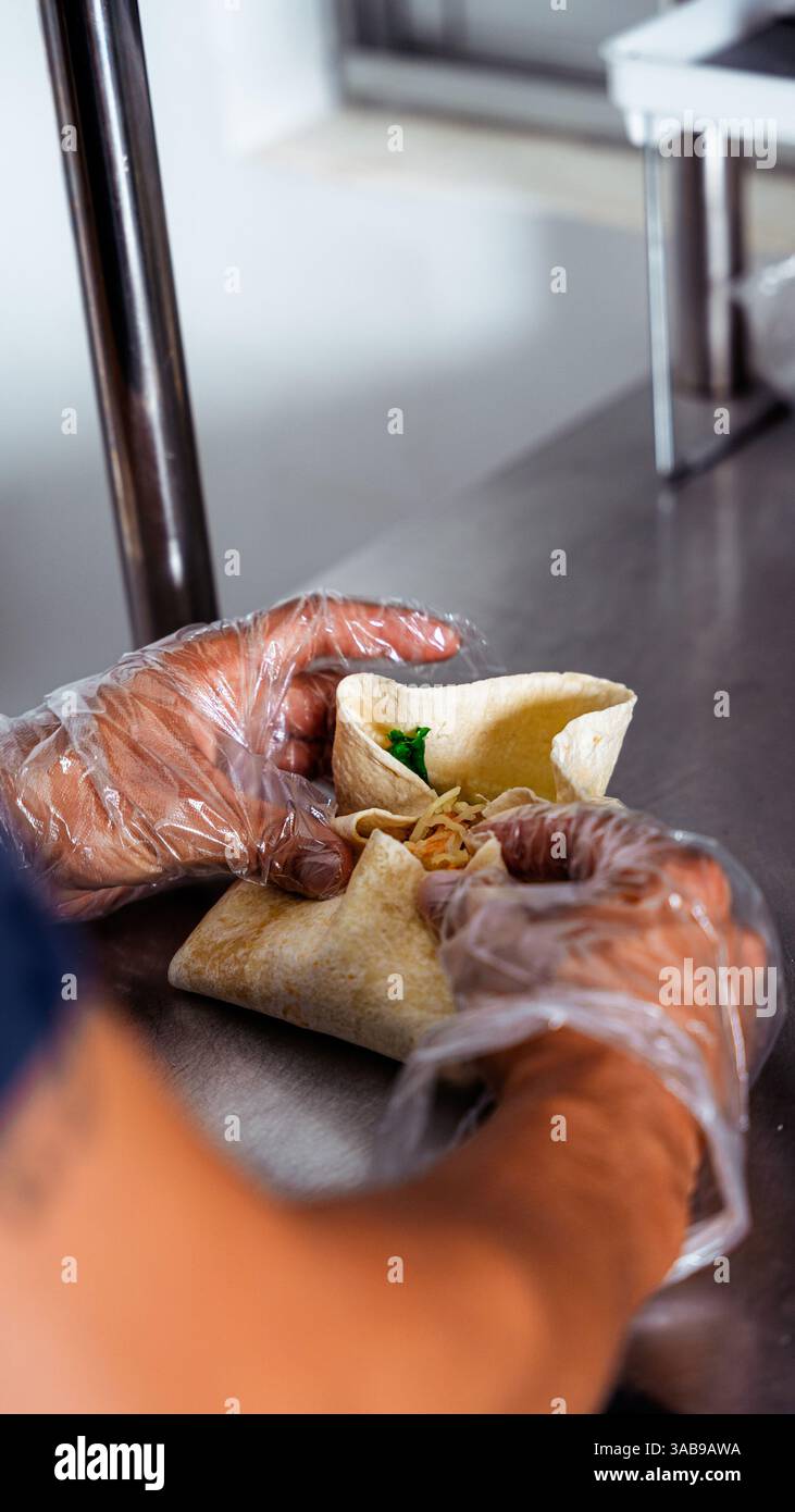 Les mains gantées assemblent habilement un enveloppement savoureux sur une surface en acier inoxydable. Cette scène capture l'essence vibrante d'une cuisine de café confortable en action. Banque D'Images