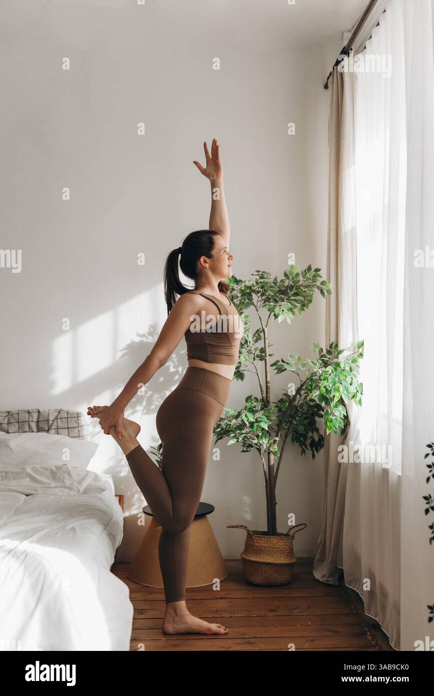 Une jeune femme pratique le yoga dans une chambre confortable et ensoleillée avec du parquet, des plantes luxuriantes et une literie moelleuse. L'atmosphère sereine favorise la pleine conscience Banque D'Images
