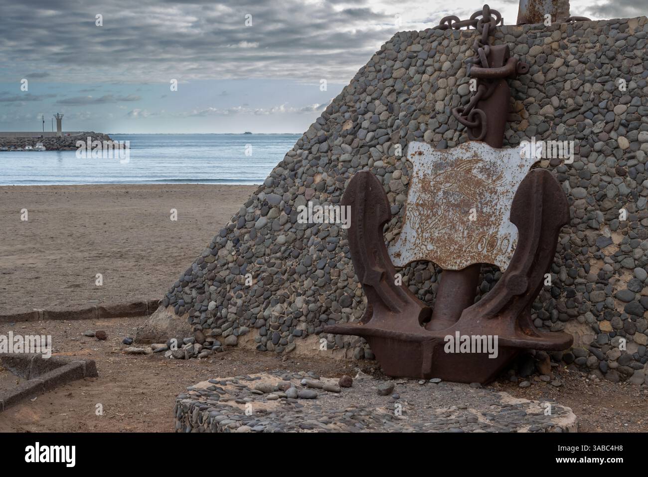 Monument couvert de cailloux et d'une ancre rouillée ancienne sur la côte de l'océan Atlantique. Jetée en arrière-plan. Ciel nocturne nuageux. Castillo del Romeral, Banque D'Images