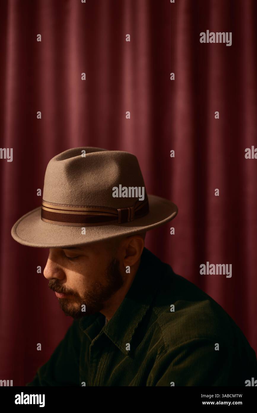 Portrait d'un homme avec une barbe portant un chapeau Fedora brun clair assis dans un studio sur un fond drapé rose. Banque D'Images