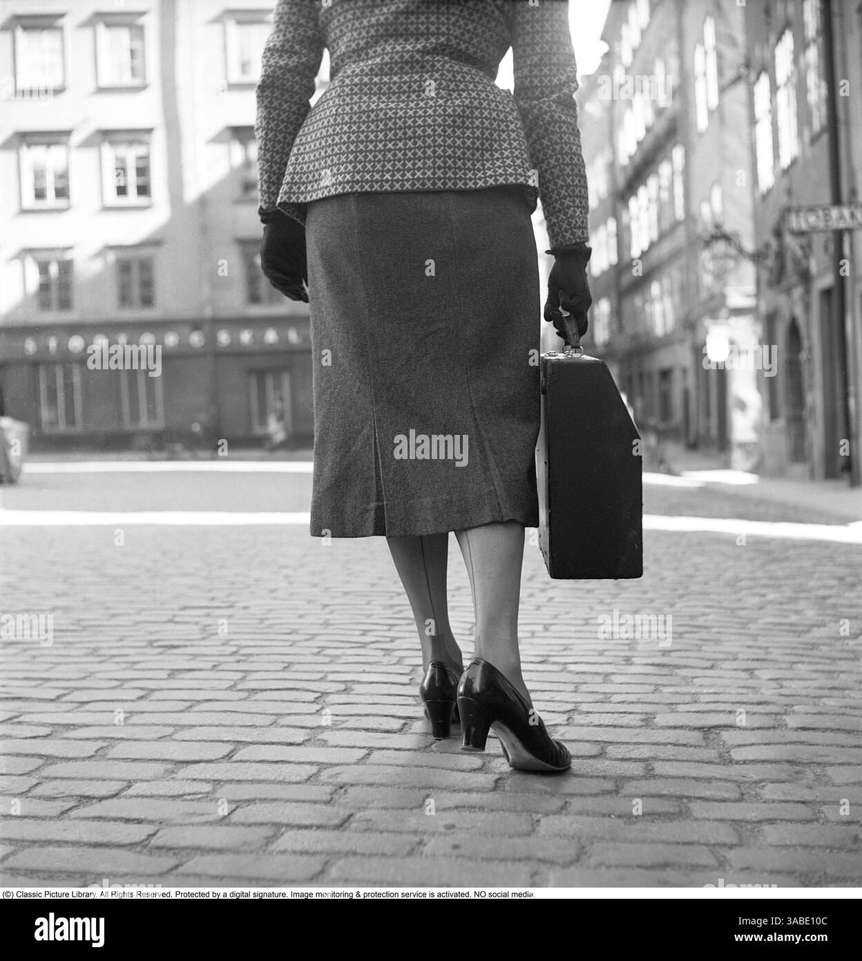 Une femme des années 1950 marchant le long d'une rue pavée à Gamla stan, Suède, en 1955. Elle est habillée élégamment d'une veste à motifs, d'une jupe longueur genou, de gants et de chaussures à talons hauts, reflétant la mode de l'époque. Dans une main, elle transporte une machine à écrire de voyage dans son étui désigné, conçu pour un transport facile. Ce modèle de machine à écrire était plus petit qu'un modèle standard, ce qui le rendait plus pratique pour les écrivains, les journalistes et les hommes d'affaires qui avaient besoin de travailler en déplacement avant l'avènement des ordinateurs portables. La photographie donne un aperçu d'une époque passée, où les machines à écrire portatives étaient essentielles Banque D'Images