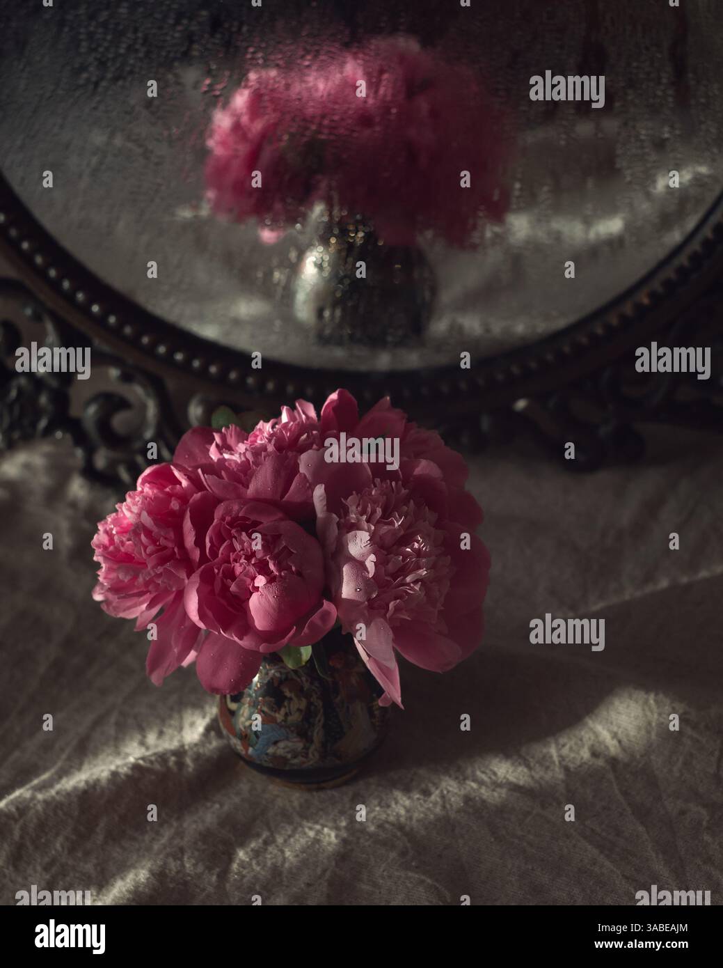 Nature morte nostalgique représentant un bouquet de pivoines roses dans un vase orné, derrière lequel se trouve un miroir antique reflétant les fleurs à travers des gouttes Banque D'Images