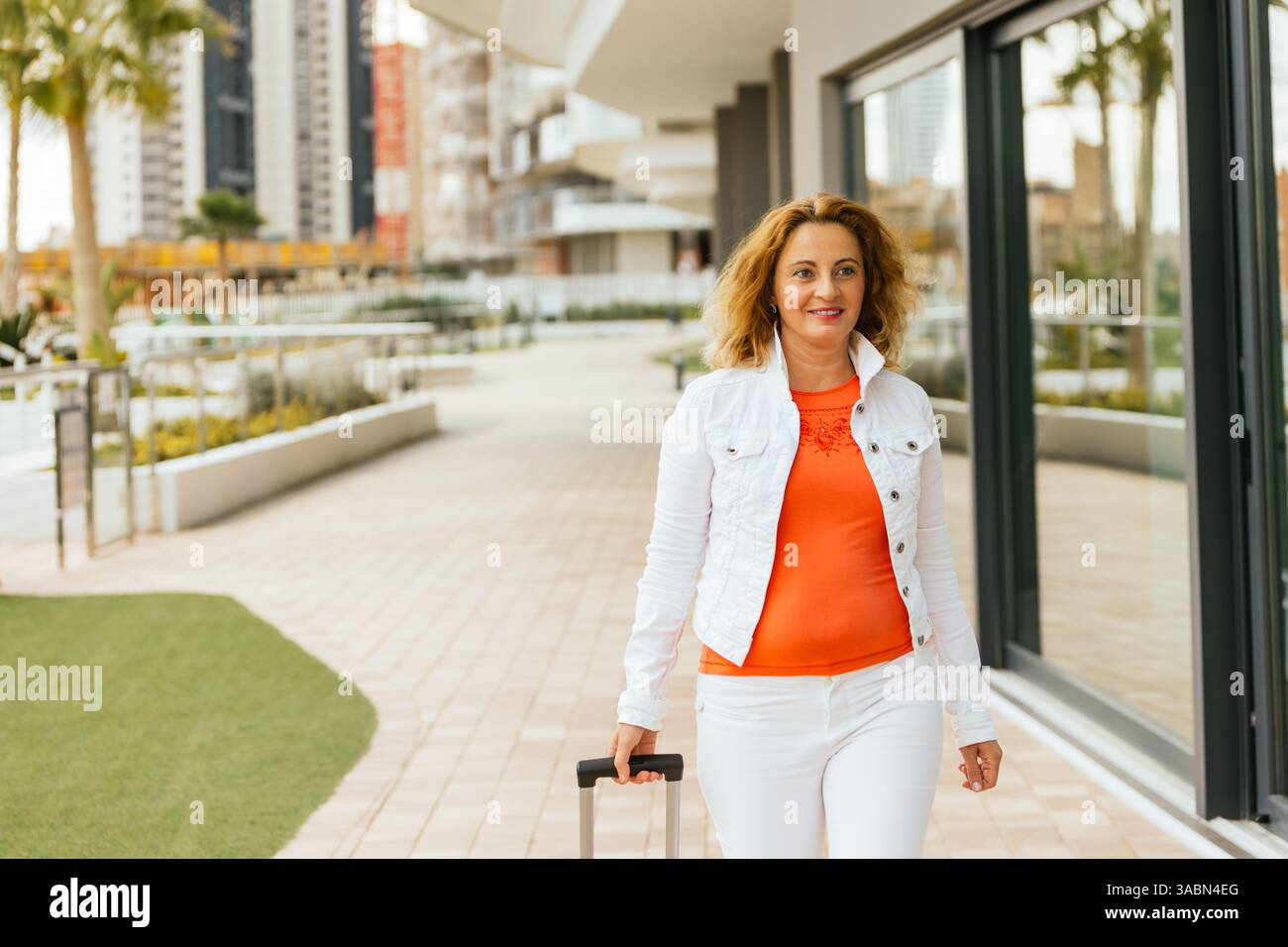 Femme d'âge moyen souriante dans un haut orange et veste blanche marchant devant les fenêtres de l'hôtel avec une valise, sur fond de bâtiments modernes A. Banque D'Images