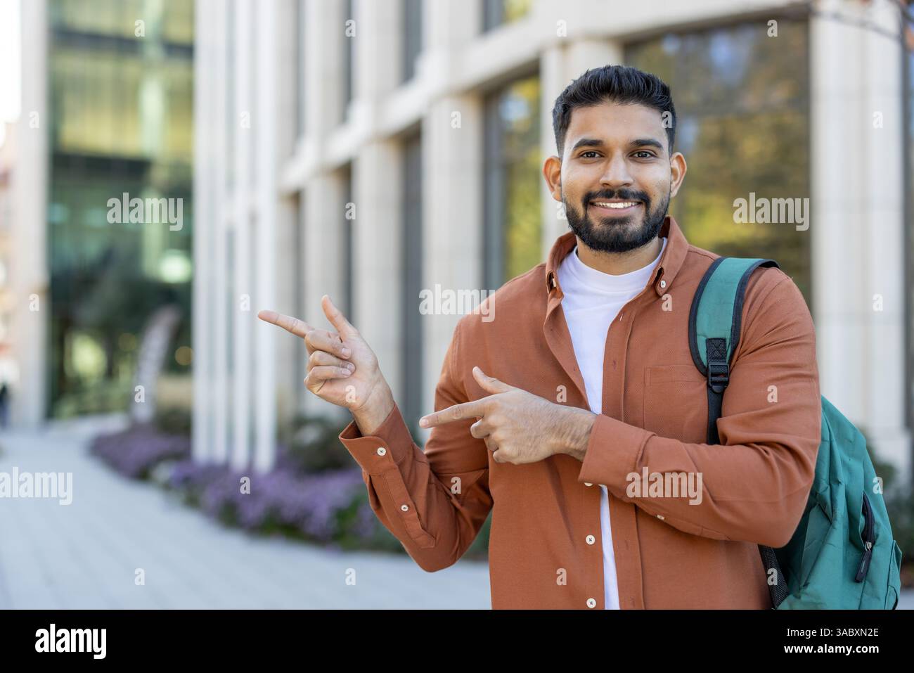 Un jeune homme souriant fait des gestes avec ses mains, pointant vers une zone d'intérêt. Il porte un sac à dos devant un bâtiment moderne. Banque D'Images